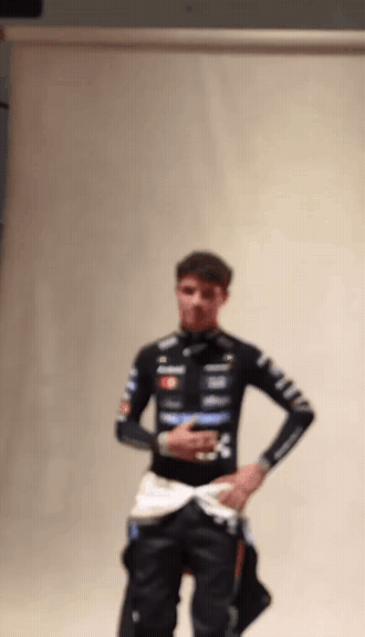 A Man In A Racing Suit Is Standing In Front Of A White Wall With His Hands In His Pockets . GIF