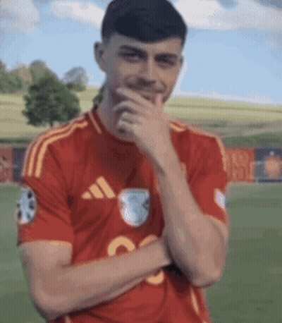 A Man In A Red Adidas Shirt Is Covering His Mouth With His Hand While Standing On A Field . GIF