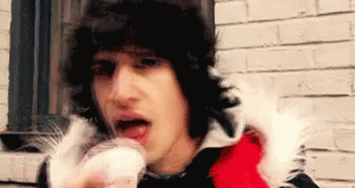 A Man In A Red And Black Jacket Is Eating A Cupcake In Front Of A Brick Wall . GIF