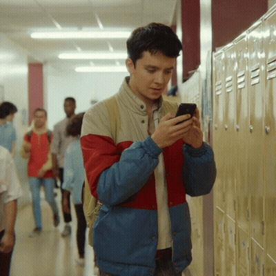 A Man In A Red And Blue Jacket Looks At His Phone In A Hallway GIF