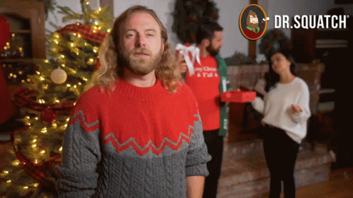 A Man In A Red And Gray Sweater Is Standing In Front Of A Christmas Tree GIF