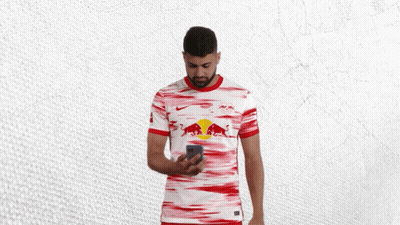 A Man In A Red And White Jersey With A Bull On It Looks At His Phone GIF