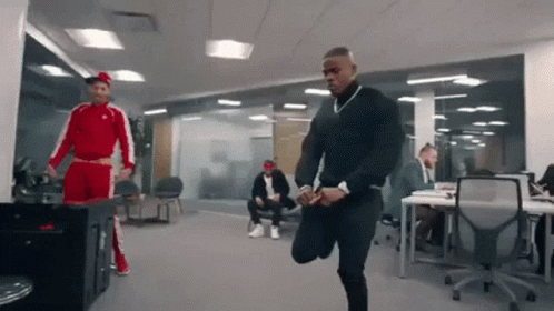 A Man In A Red And White Tracksuit Is Stretching His Legs In An Office While Other Men Sit At Desks . GIF