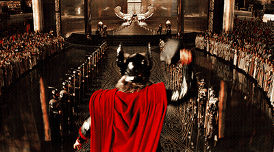 A Man In A Red Cape Is Holding A Hammer In Front Of A Large Crowd GIF