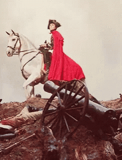 A Man In A Red Cape Is Riding A White Horse Next To A Cannon GIF