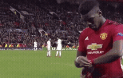 A Man In A Red Chevrolet Jersey Is Standing On A Soccer Field Looking At His Watch . GIF