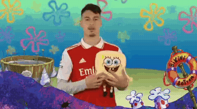 A Man In A Red Emirates Jersey Holds A Stuffed Spongebob Doll GIF