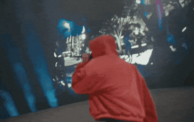A Man In A Red Hoodie Is Singing Into A Microphone In Front Of A Crowd GIF