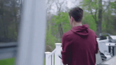 A Man In A Red Hoodie Is Standing On A Balcony Looking Out A Window . GIF