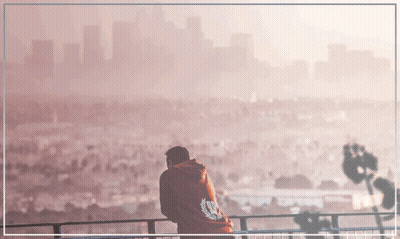 A Man In A Red Hoodie Stands On A Balcony With A City In The Background GIF