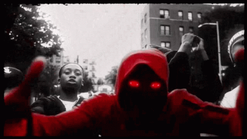 A Man In A Red Hoodie With Red Eyes Stands In A Crowd Of People GIF