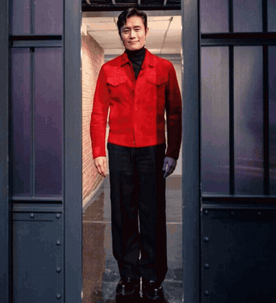 A Man In A Red Jacket And Black Pants Stands In A Dark Room GIF