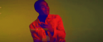 A Man In A Red Jacket And Black Turtleneck Is Dancing In Front Of A Yellow Wall . GIF