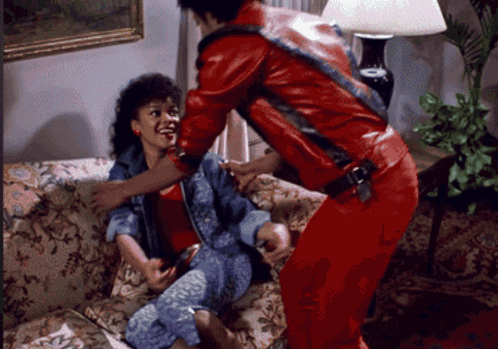 A Man In A Red Jacket Holds A Woman On A Couch GIF