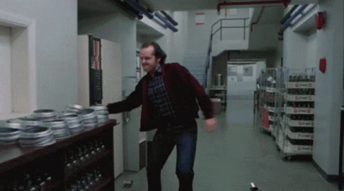 A Man In A Red Jacket Is Dancing In A Hallway With A Shelf Full Of Reels Of Film GIF