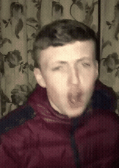 A Man In A Red Jacket Is Making A Funny Face In Front Of A Curtain With Flowers On It GIF