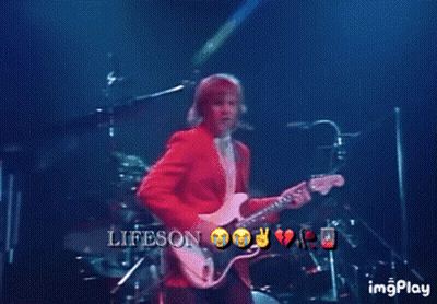 A Man In A Red Jacket Is Playing A Guitar On A Stage With The Words Lifeson On The Bottom Right GIF