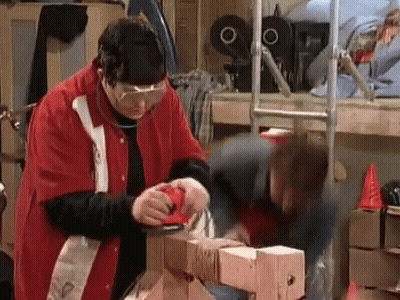 A Man In A Red Jacket Is Sanding A Piece Of Wood While A Woman Watches . GIF