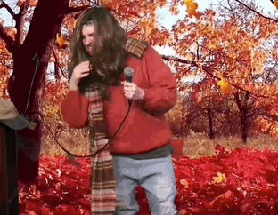 A Man In A Red Jacket Is Singing Into A Microphone In A Field Of Leaves . GIF