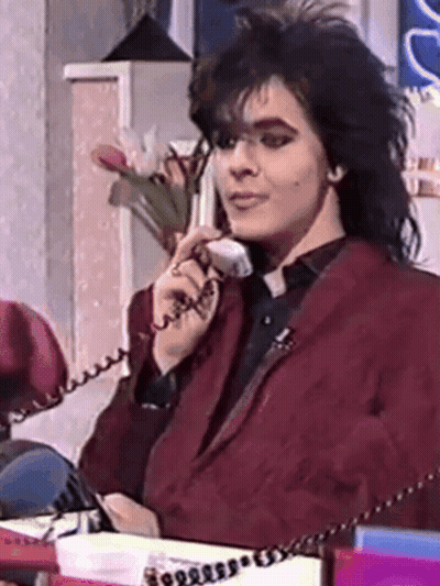 A Man In A Red Jacket Is Sitting At A Desk Talking On A Phone GIF