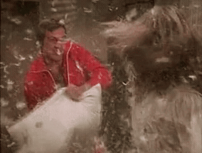 A Man In A Red Jacket Is Sitting In A Pile Of Feathers While A Woman Throws Feathers At Him . GIF