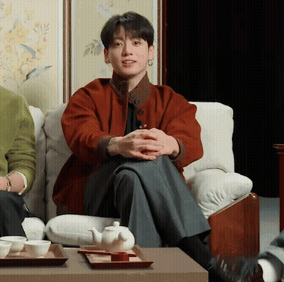 A Man In A Red Jacket Is Sitting On A Couch With His Legs Crossed GIF