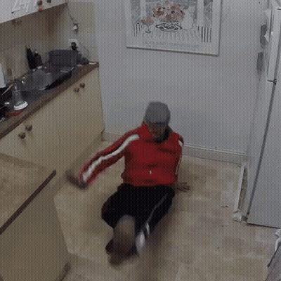 A Man In A Red Jacket Is Sitting On The Floor In A Kitchen With A Picture On The Wall Above Him . GIF