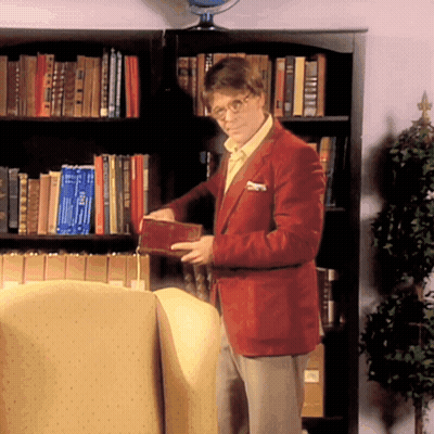 A Man In A Red Jacket Is Standing In Front Of A Bookshelf Holding A Book That Says ' I Love You ' GIF