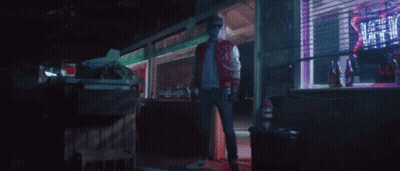A Man In A Red Jacket Is Standing In Front Of A Store With A Neon Sign That Says ' Hot ' On It . GIF