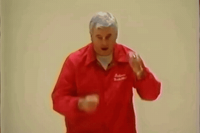 A Man In A Red Jacket Is Standing In Front Of A White Wall And Making A Funny Face . GIF