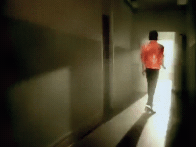 A Man In A Red Jacket Is Walking Down A Dark Hallway Towards A Light . GIF