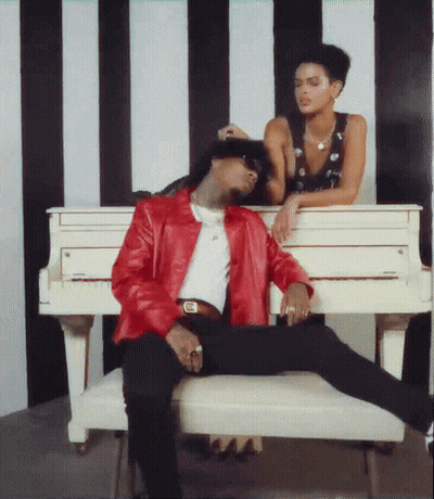 A Man In A Red Jacket Sits On A Bench Next To A Woman Leaning On A White Piano GIF