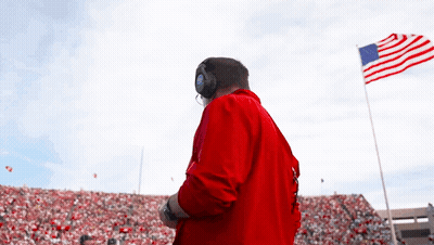 A Man In A Red Jacket Stands In Front Of A Large American Flag GIF