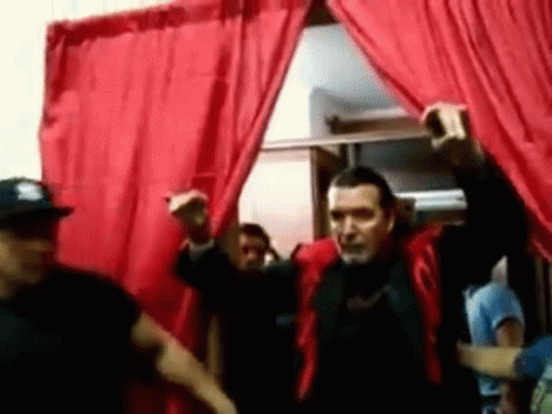 A Man In A Red Jacket Stands In Front Of A Red Curtain With His Arms In The Air GIF