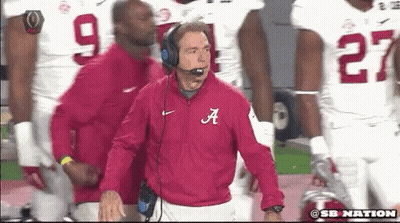 A Man In A Red Jacket With The Letter A On It Stands In Front Of A Group Of Football Players GIF