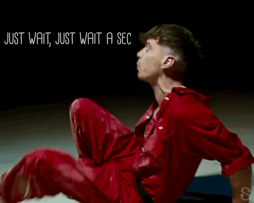 A Man In A Red Jumpsuit Is Sitting On The Floor With The Words Just Wait Just Wait A Sec Written Above Him GIF