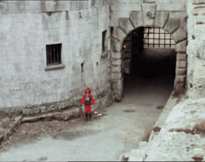 A Man In A Red Knight Costume Stands In Front Of A Stone Building GIF