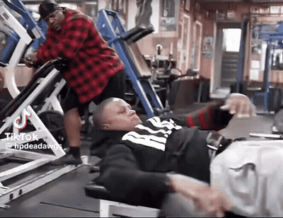 A Man In A Red Plaid Shirt Is Standing Next To A Man In A Black And White Shirt On A Leg Press Machine . GIF