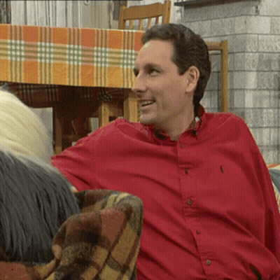 A Man In A Red Polo Shirt Smiles While Sitting On A Couch GIF