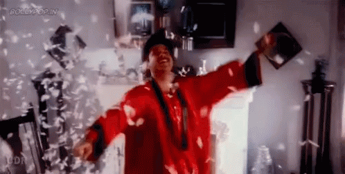 A Man In A Red Robe Is Standing In A Room With Feathers Falling Around Him . GIF