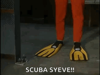 A Man In A Red Scuba Suit And Yellow Gloves Is Standing In A Room . GIF