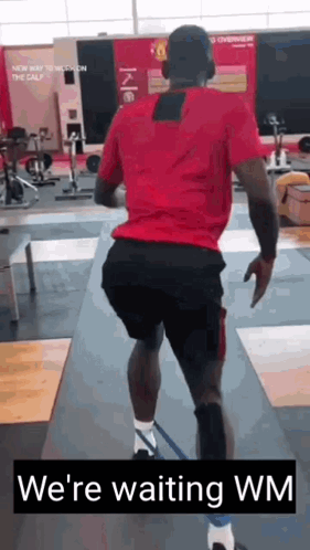 A Man In A Red Shirt And Black Shorts Is Running In A Gym With The Words `` We 'Re Waiting Wm '' . GIF