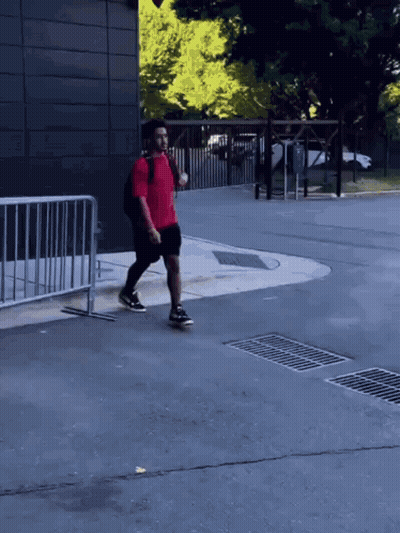 A Man In A Red Shirt And Black Shorts Is Walking Down A Street GIF