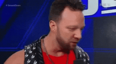 A Man In A Red Shirt And Black Vest Is Talking Into A Microphone On A Stage . GIF