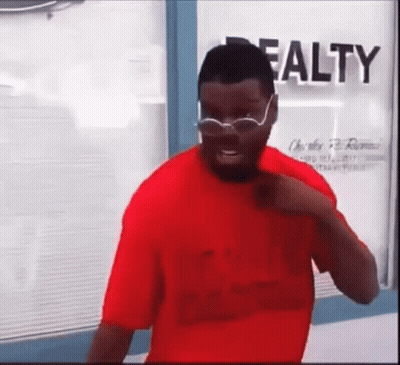 A Man In A Red Shirt And Glasses Is Standing In Front Of A Window . GIF