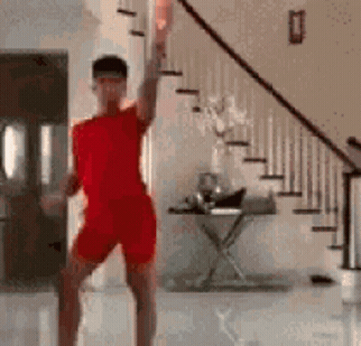 A Man In A Red Shirt And Shorts Is Dancing In A Living Room Next To Stairs . GIF