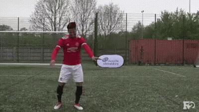 A Man In A Red Shirt And White Shorts Is Holding A Head & Shoulders Sign On A Soccer Field . GIF
