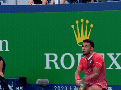 A Man In A Red Shirt Holds A Tennis Racquet In Front Of A Rolex Sign GIF