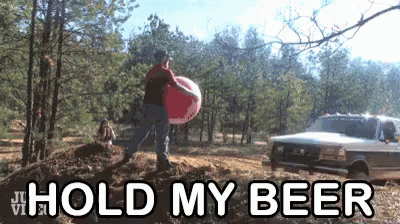 A Man In A Red Shirt Is Carrying A Large Inflatable Ball With The Words Hold My Beer Behind Him GIF
