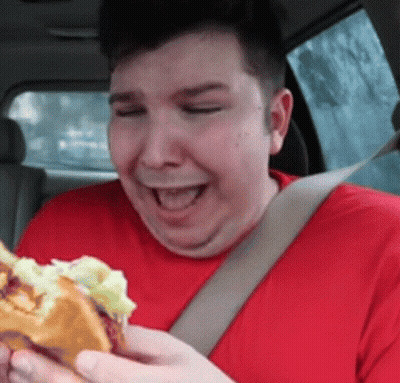 A Man In A Red Shirt Is Eating A Hamburger While Wearing A Seat Belt GIF
